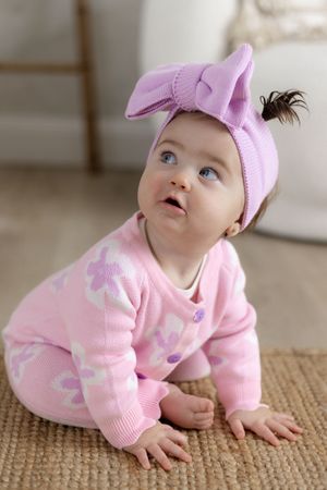 Adorable baby girl in pink floral outfit and purple bow headband sitting on a rug, looking up curiously.