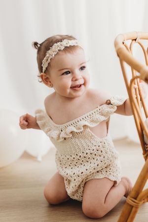 Smiling baby girl in cream crochet romper and floral headband sitting on floor next to rattan chair.