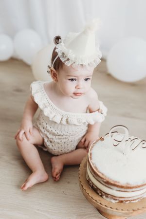 Adorable baby girl in cream knit romper and party hat sits beside first birthday cake on wooden floor.