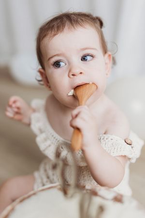 Adorable baby girl with blue eyes chewing on a wooden spoon, wearing a cream knit romper.