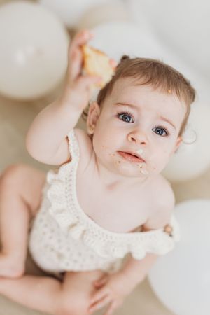 Cute baby girl with blue eyes eating cake at her first birthday party, surrounded by white balloons.