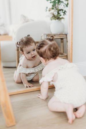 Adorable baby girl in cream romper crawling toward a floor mirror, discovering her reflection in a bright white room.