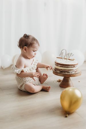 Baby girl in a polka dot romper reaching toward a naked first birthday cake with a gold “One” topper and balloon.
