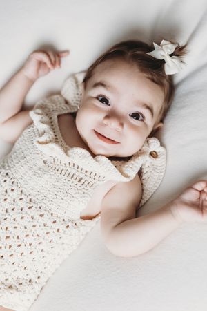 Baby wearing white crochet outfit lying on white bedding with delicate hair bows.