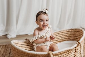 Baby sitting in woven basket wearing white lace outfit against light background.