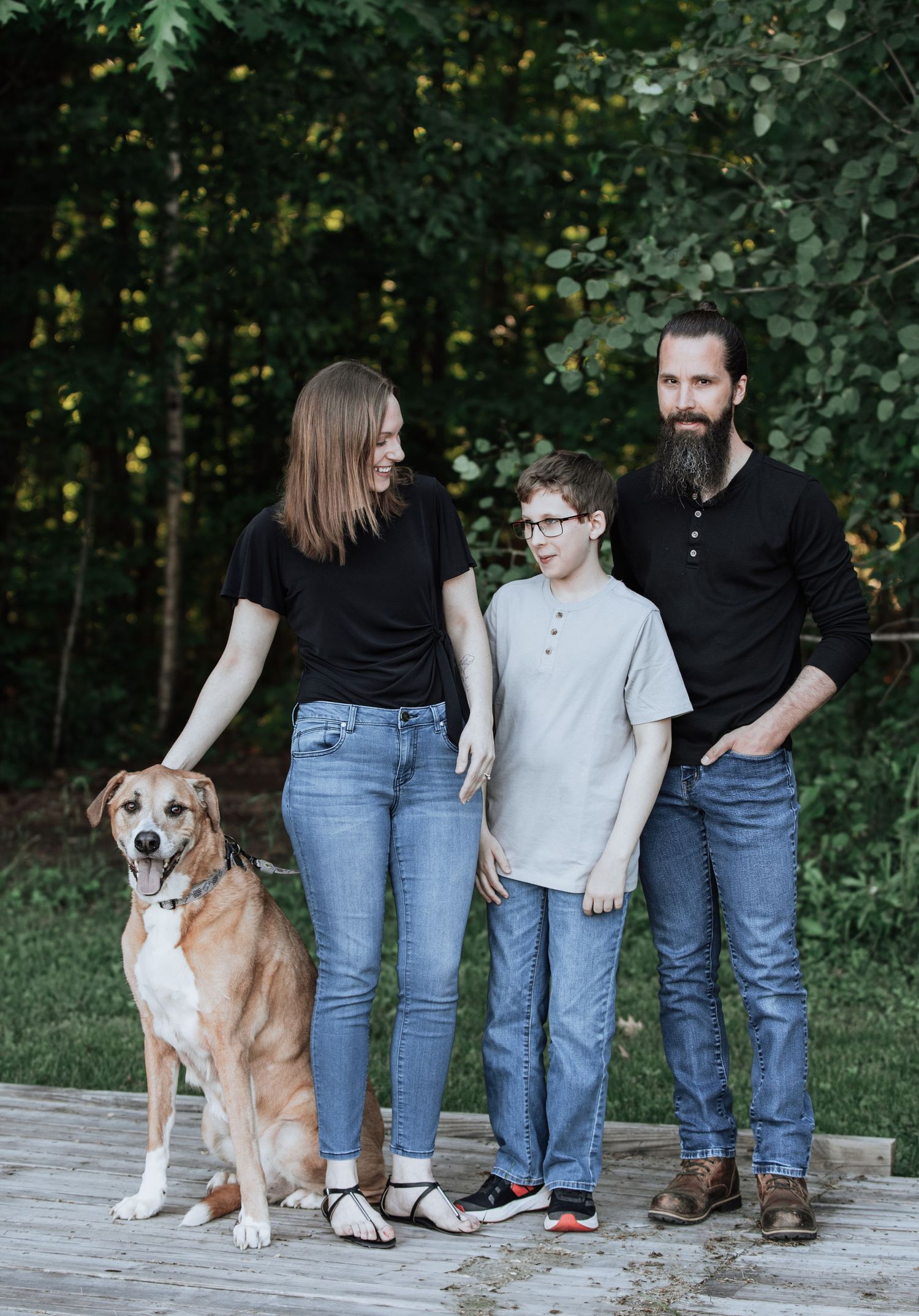 Family and their beloved dog pose together on a wooden deck near a wooded area.