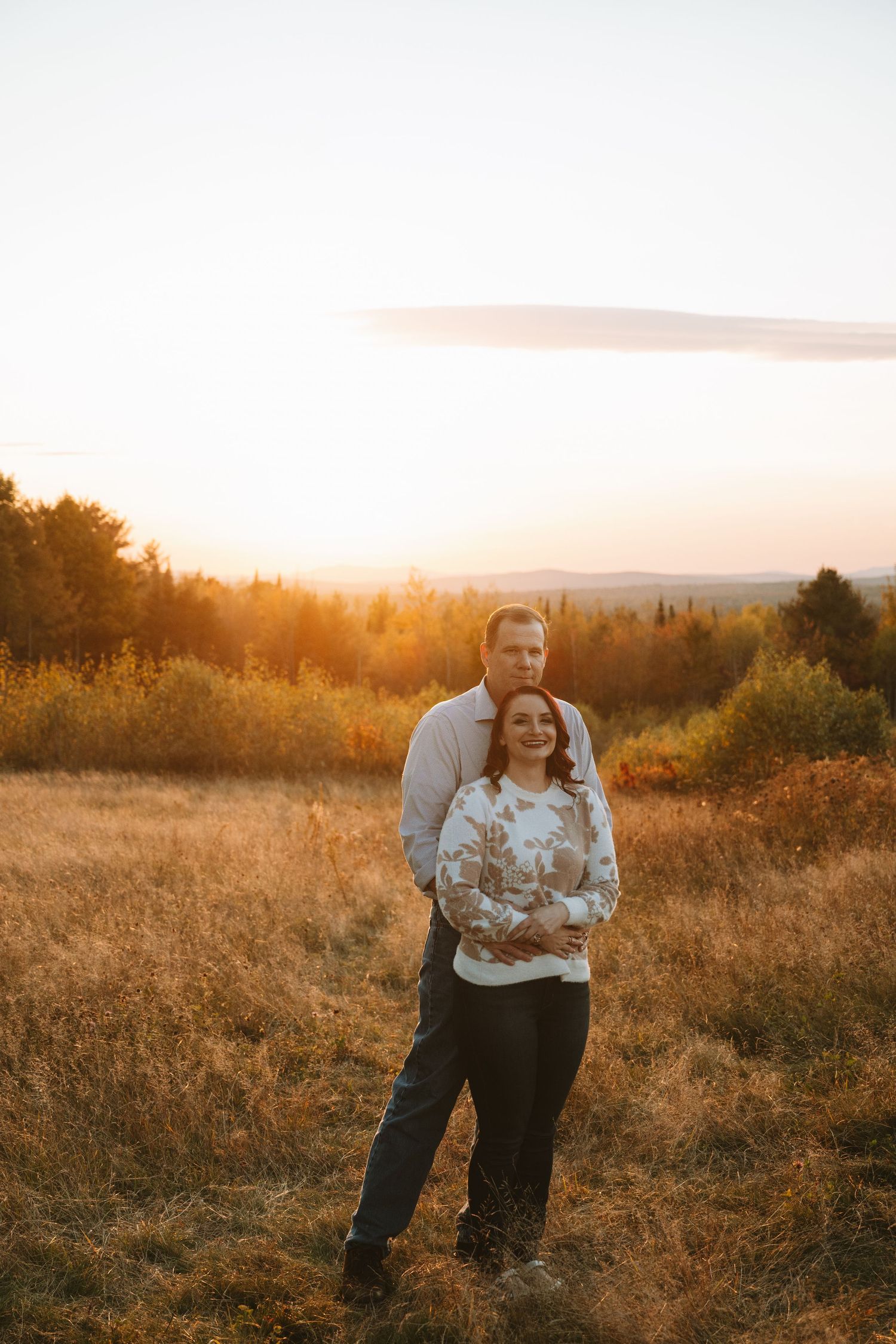 A couple embracing in a golden field during sunset with mountains in the background.