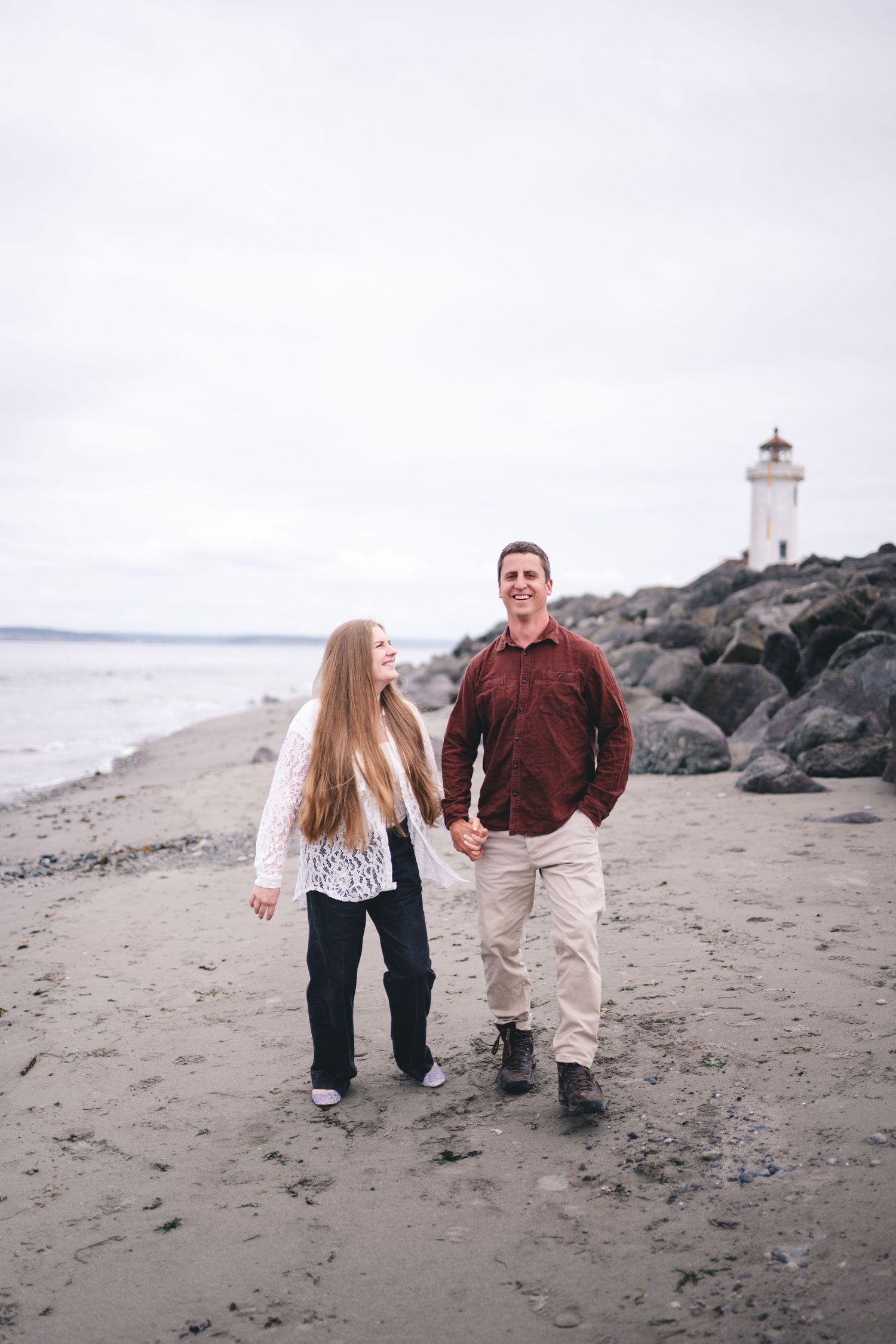 A couple walks along a beach with a lighthouse visible on rocky shore in the background.