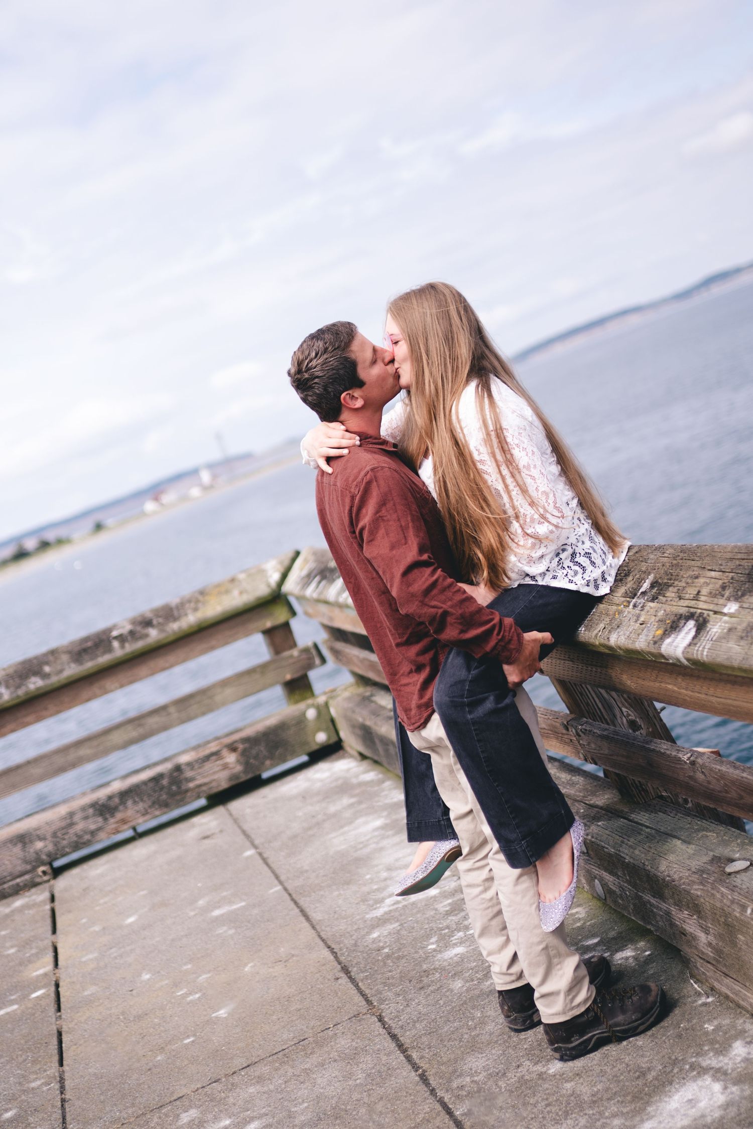 A couple embraces and kisses on a pier overlooking the ocean on a sunny day.