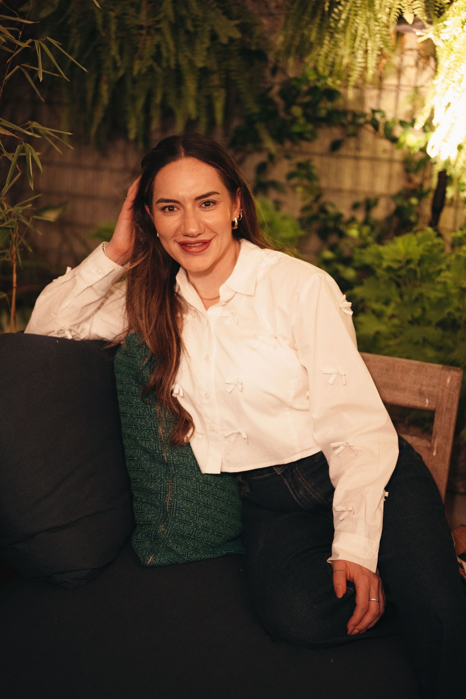 Woman in white blouse and dark outfit poses on outdoor furniture at night.