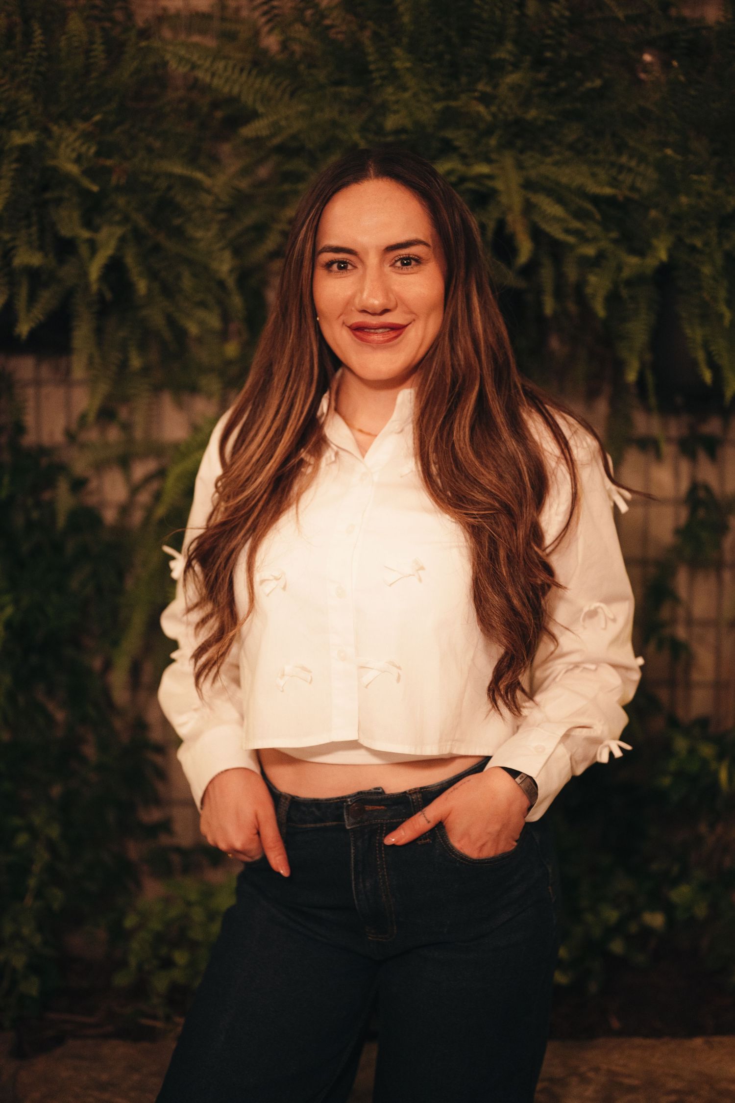 Woman in white crop top and dark pants poses against garden backdrop with evening lighting.