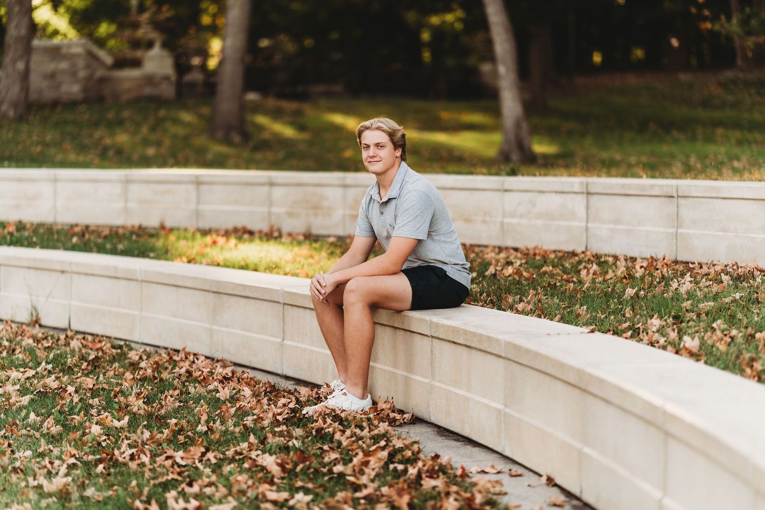 Someone in casual attire sits on a curved stone wall surrounded by autumn leaves in a park setting.