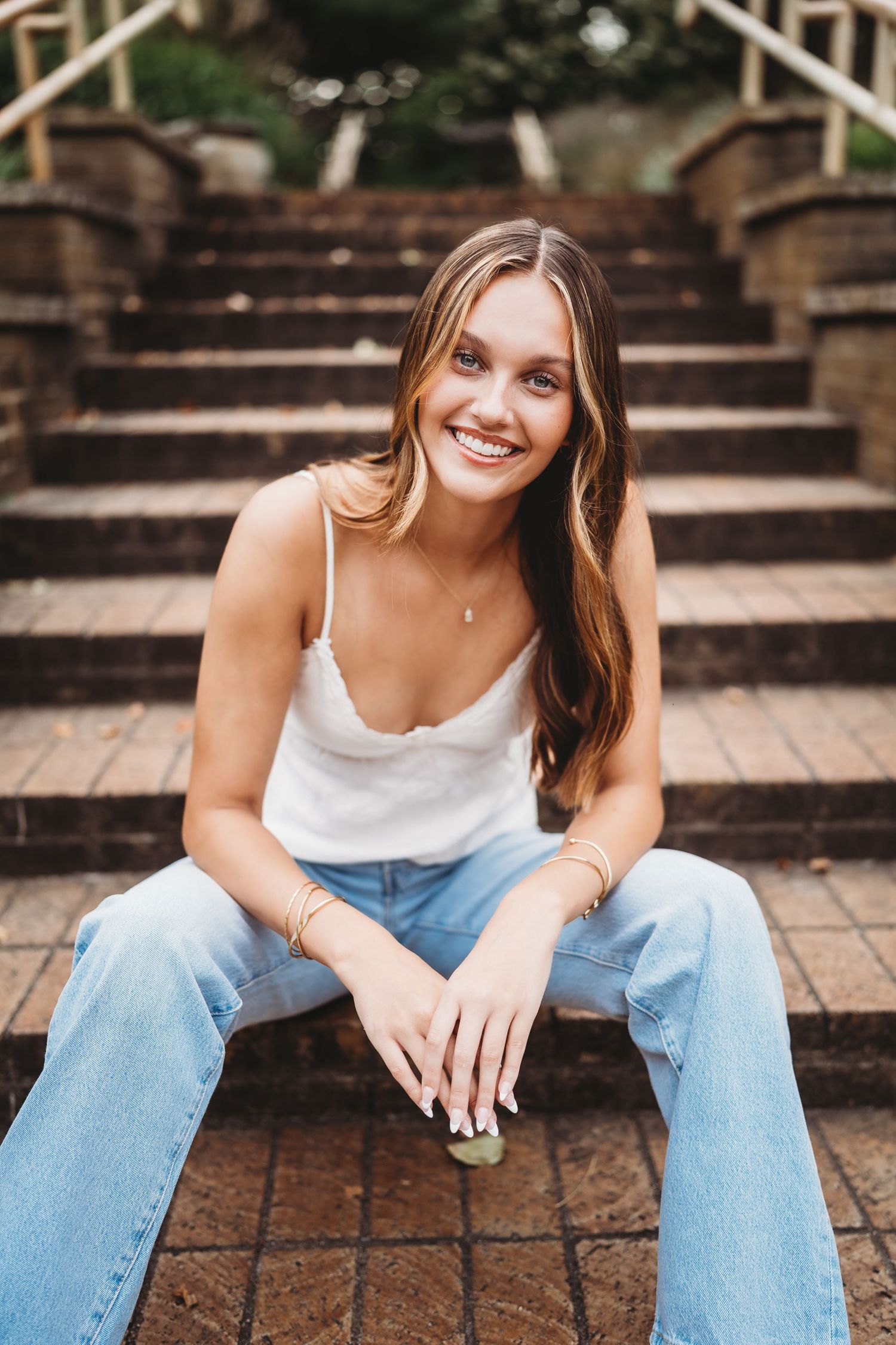 A person in a white tank top and light blue jeans sits casually on outdoor brick steps.