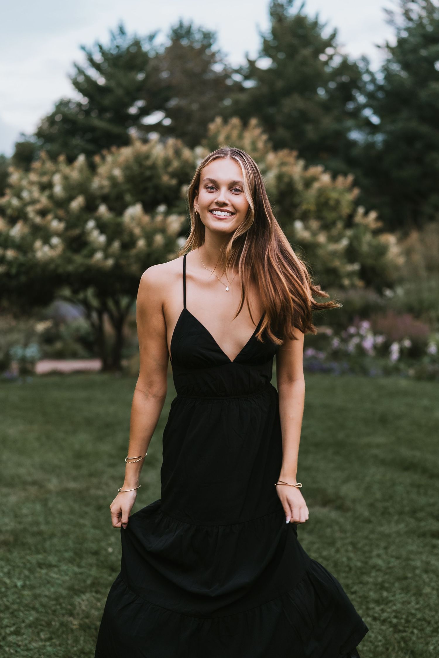 A joyful person in a black dress twirls and dances on green grass in a park setting during golden hour.
