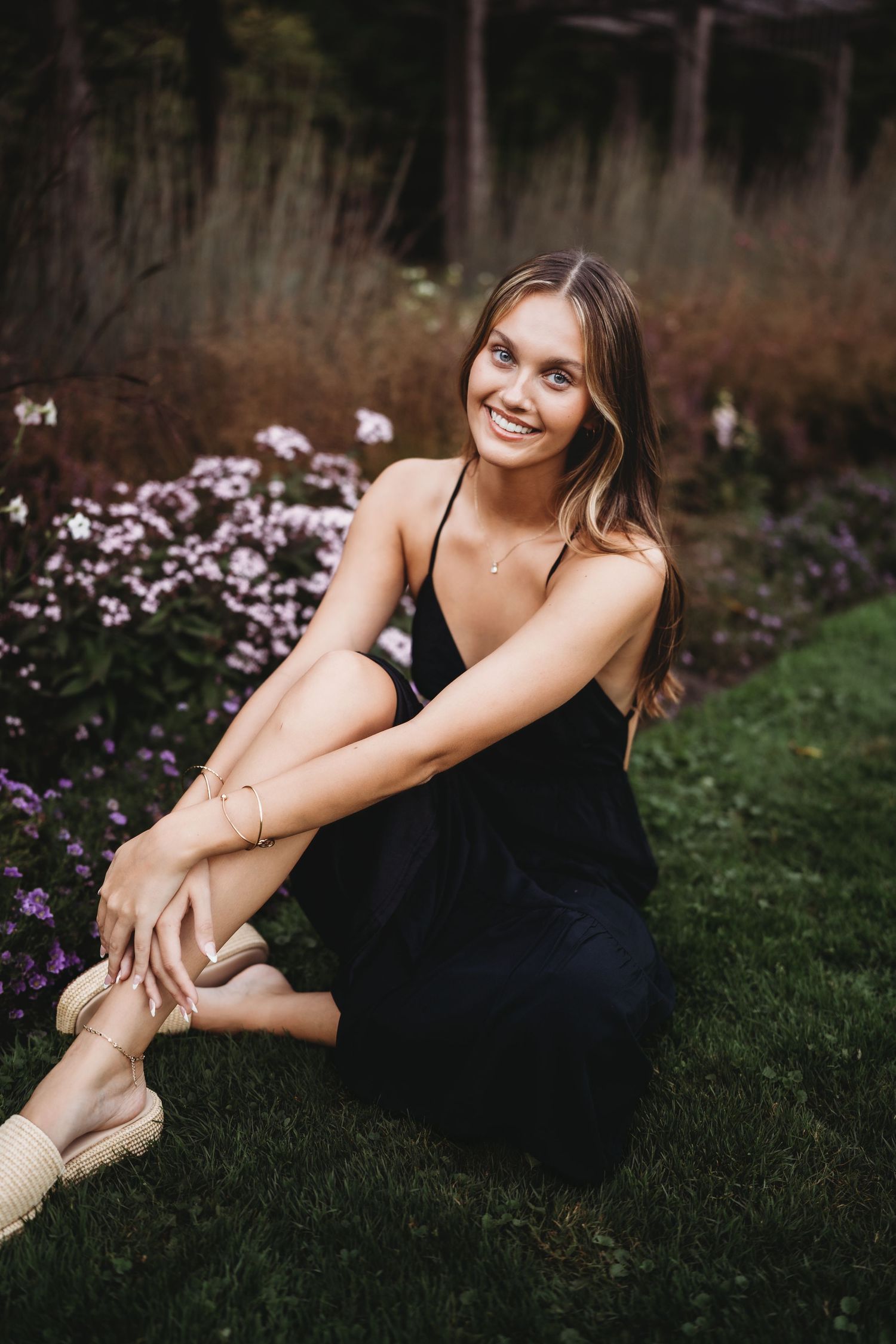 A woman in a black dress poses gracefully on grass near purple wildflowers at sunset.
