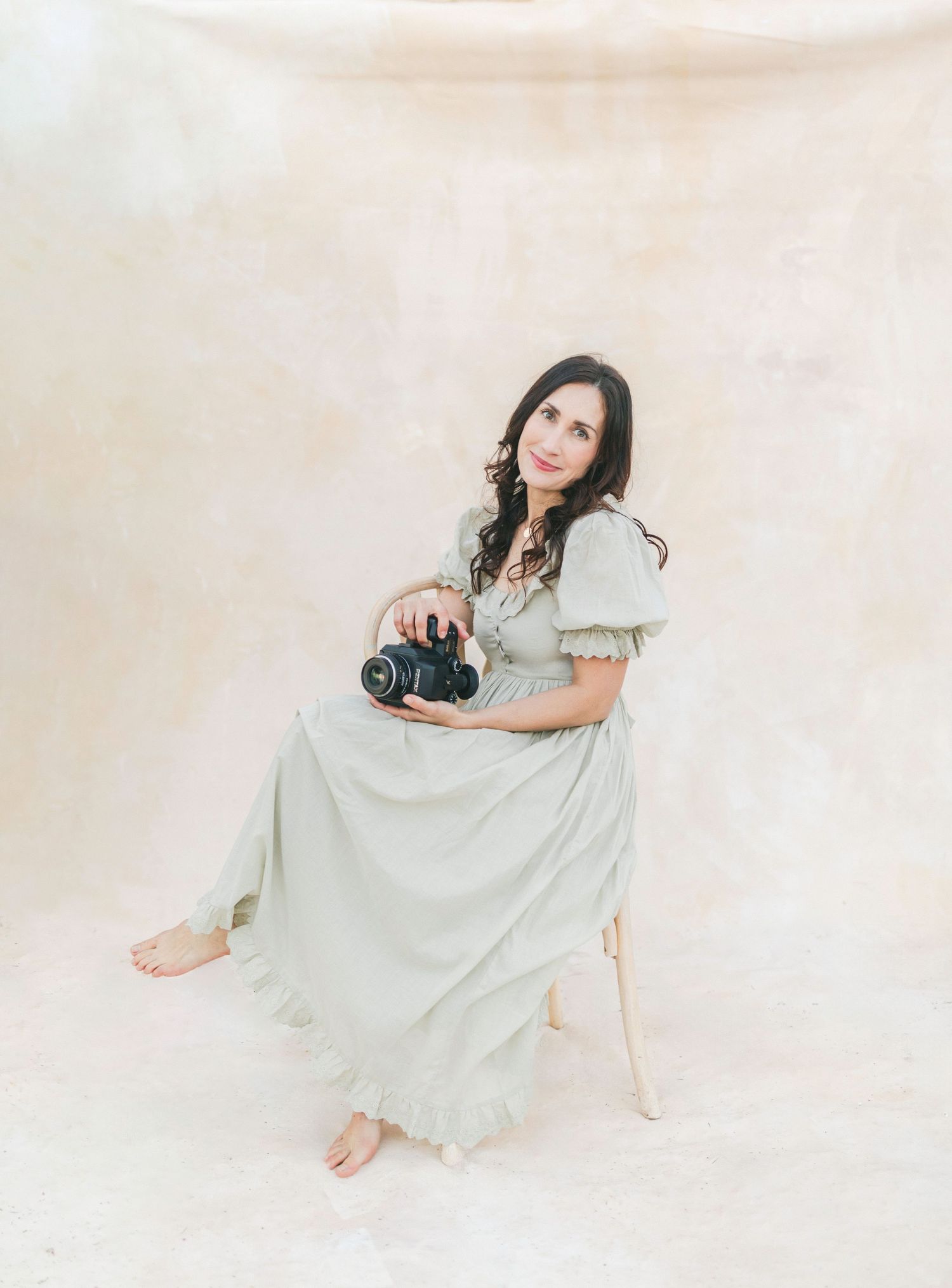 Chelsee Rawe photographer in a green vintage dress sits barefoot on a stool holding a vintage camera against a light background.