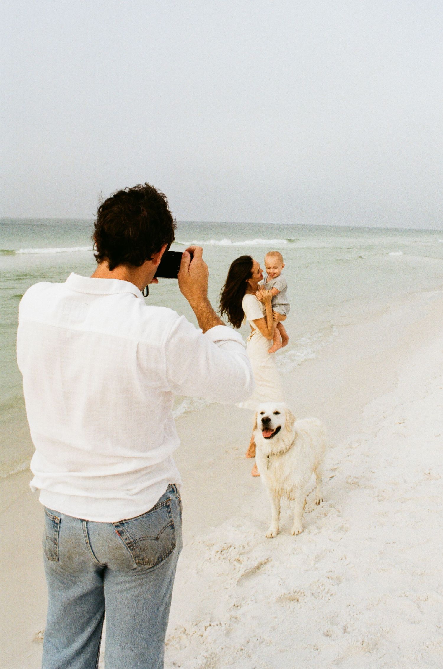 Person taking photographs on a white sandy beach with a dog nearby.