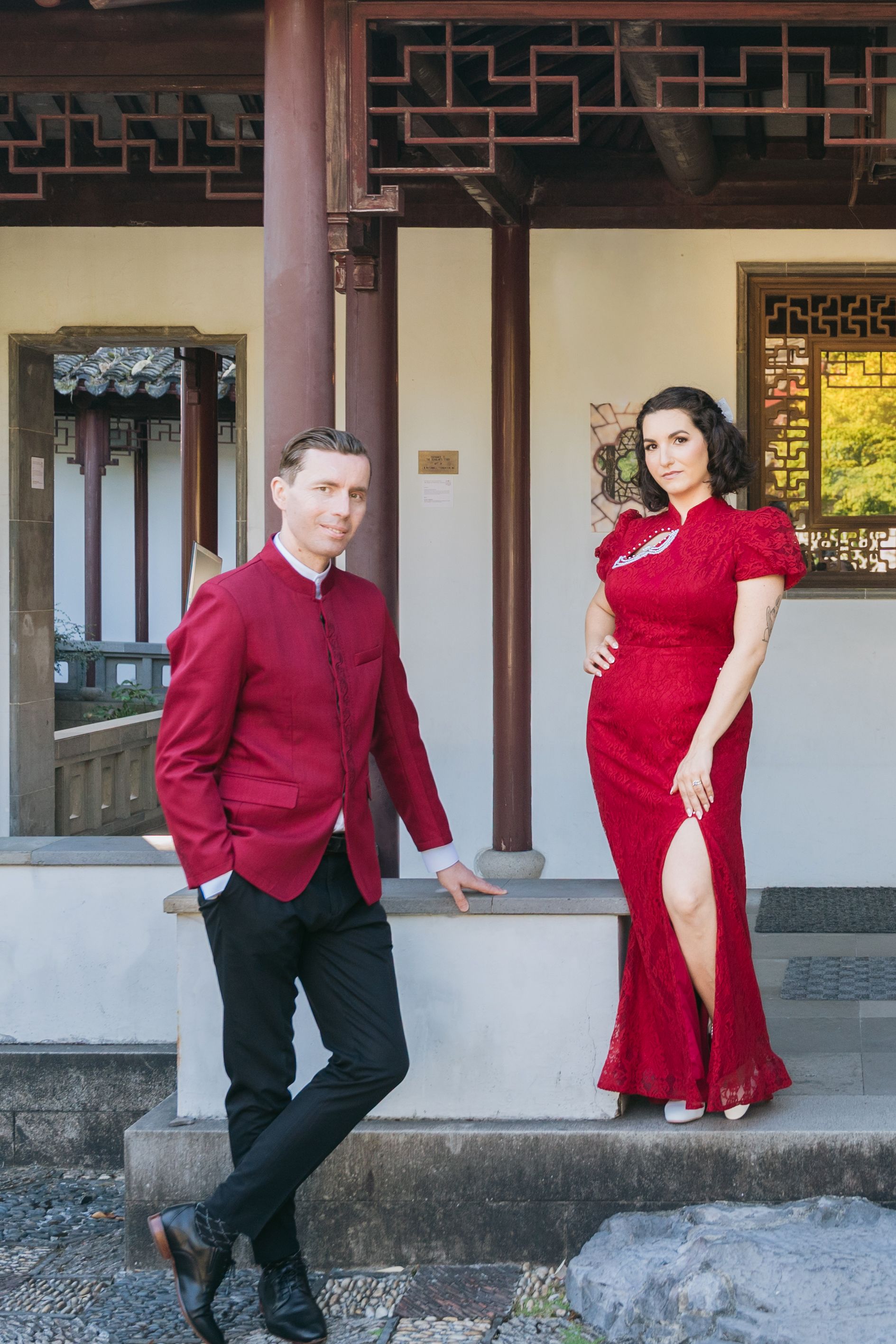A couple in red and black formal attire poses along an ornate Asian-style building.