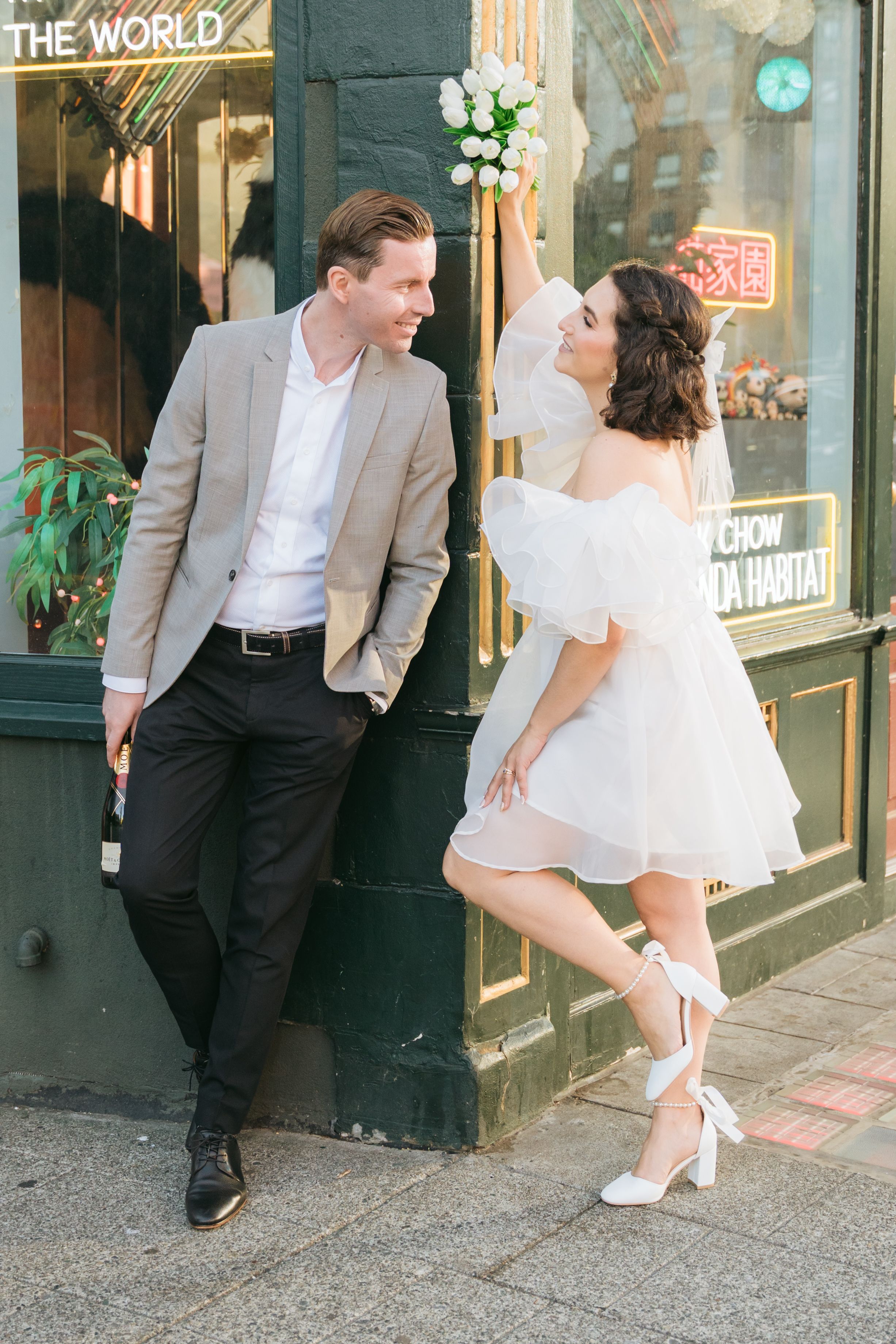 A couple in formal wear poses playfully outside a storefront with vintage signage.