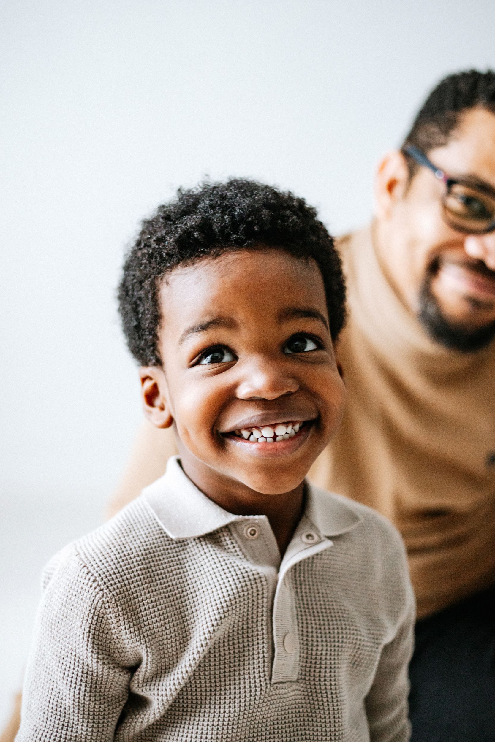 A young child wearing a beige polo shirt smiles brightly at the camera while someone sits behind them.