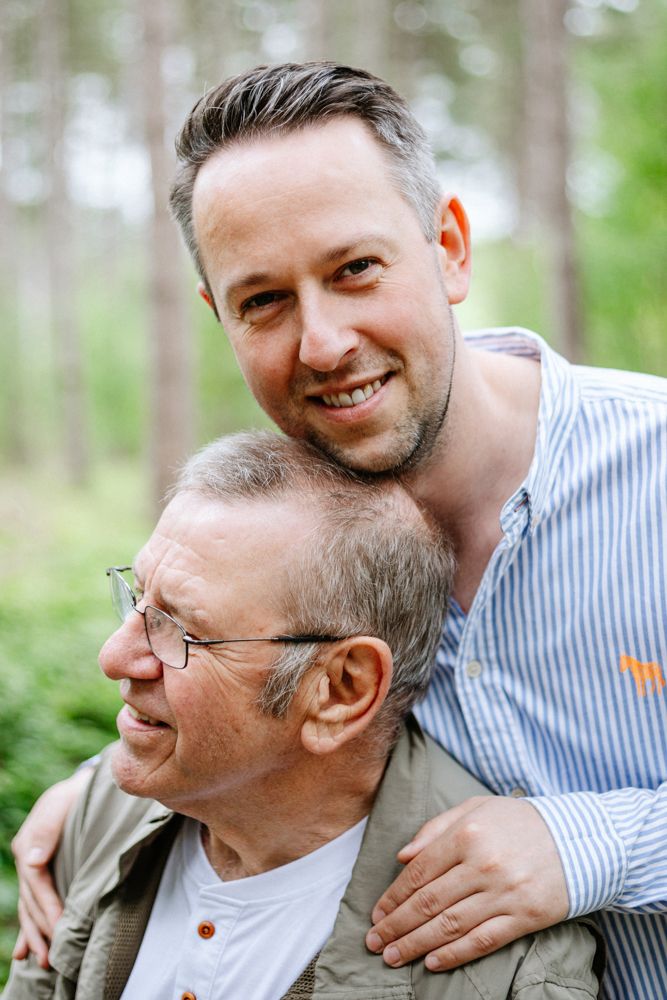 Two people share a joyful moment outdoors in a forest setting, with one wearing a striped shirt and another in casual attire.