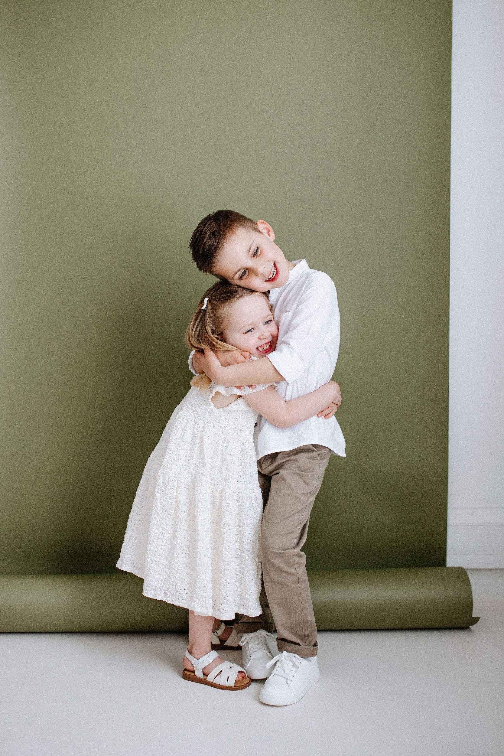 A series of four minimalist studio portraits against a sage green wall showing playful poses in white and tan clothing.
