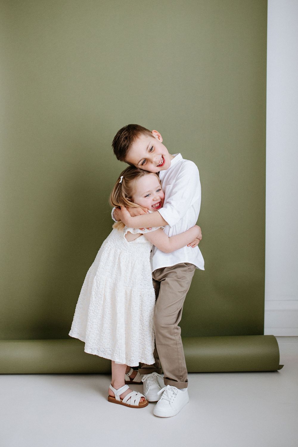 A series of four minimalist studio portraits against a sage green wall showing playful poses in white and tan clothing.