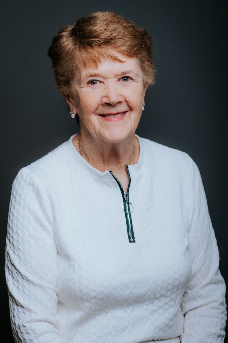 Portrait of an elderly woman smiling warmly, wearing a white textured zip-up sweater and pearl earrings, posed against a grey background