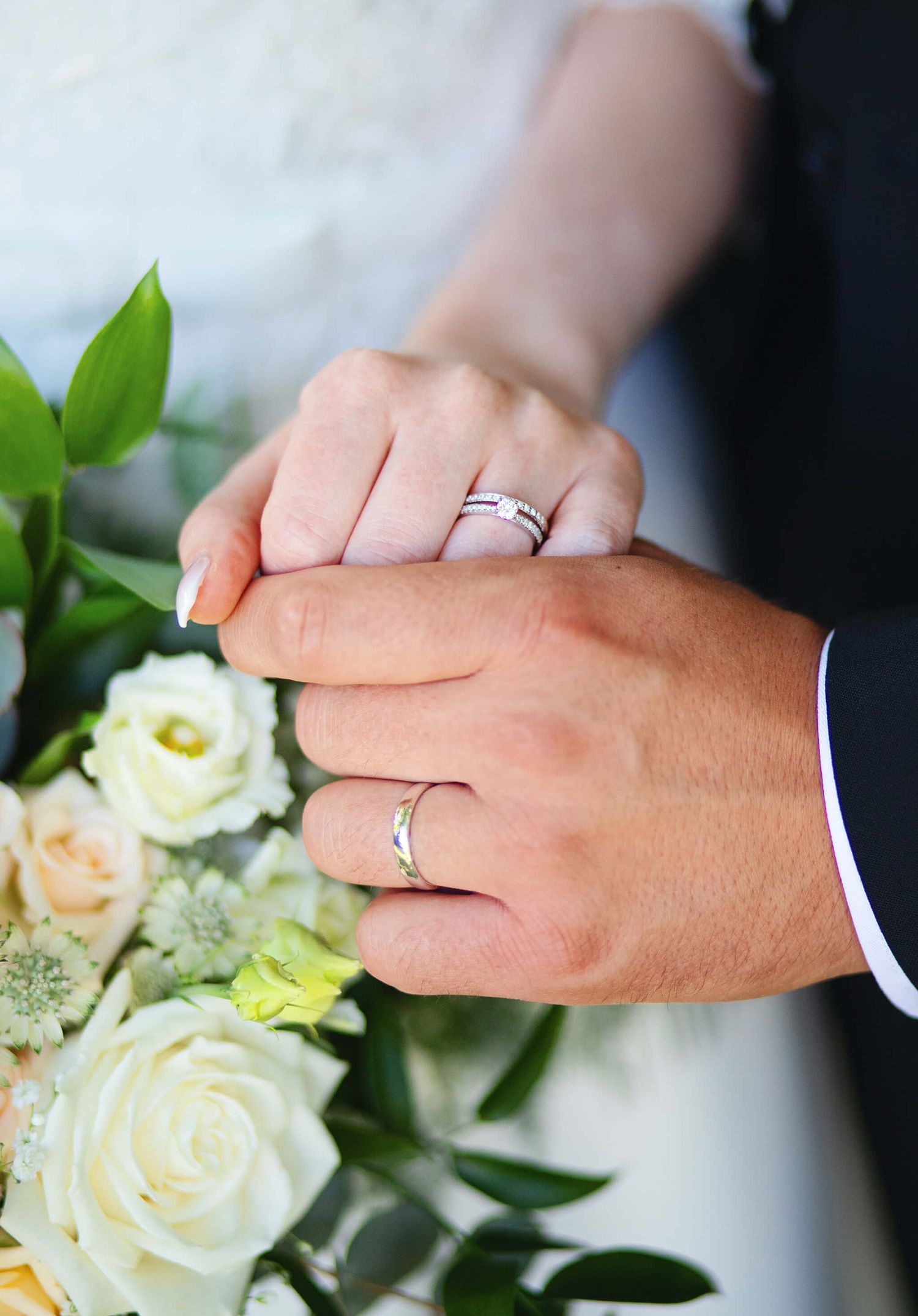 Close-up of newlyweds holding hands displaying matching wedding rings beside a bouquet of white roses.