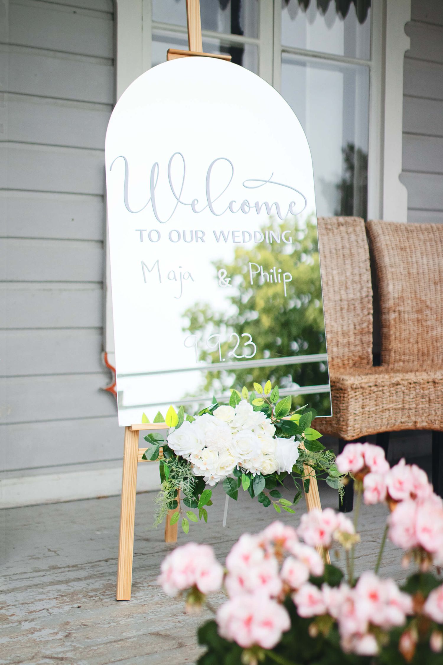 Elegant wedding welcome sign on easel with white floral arrangement and pink flowers in foreground.