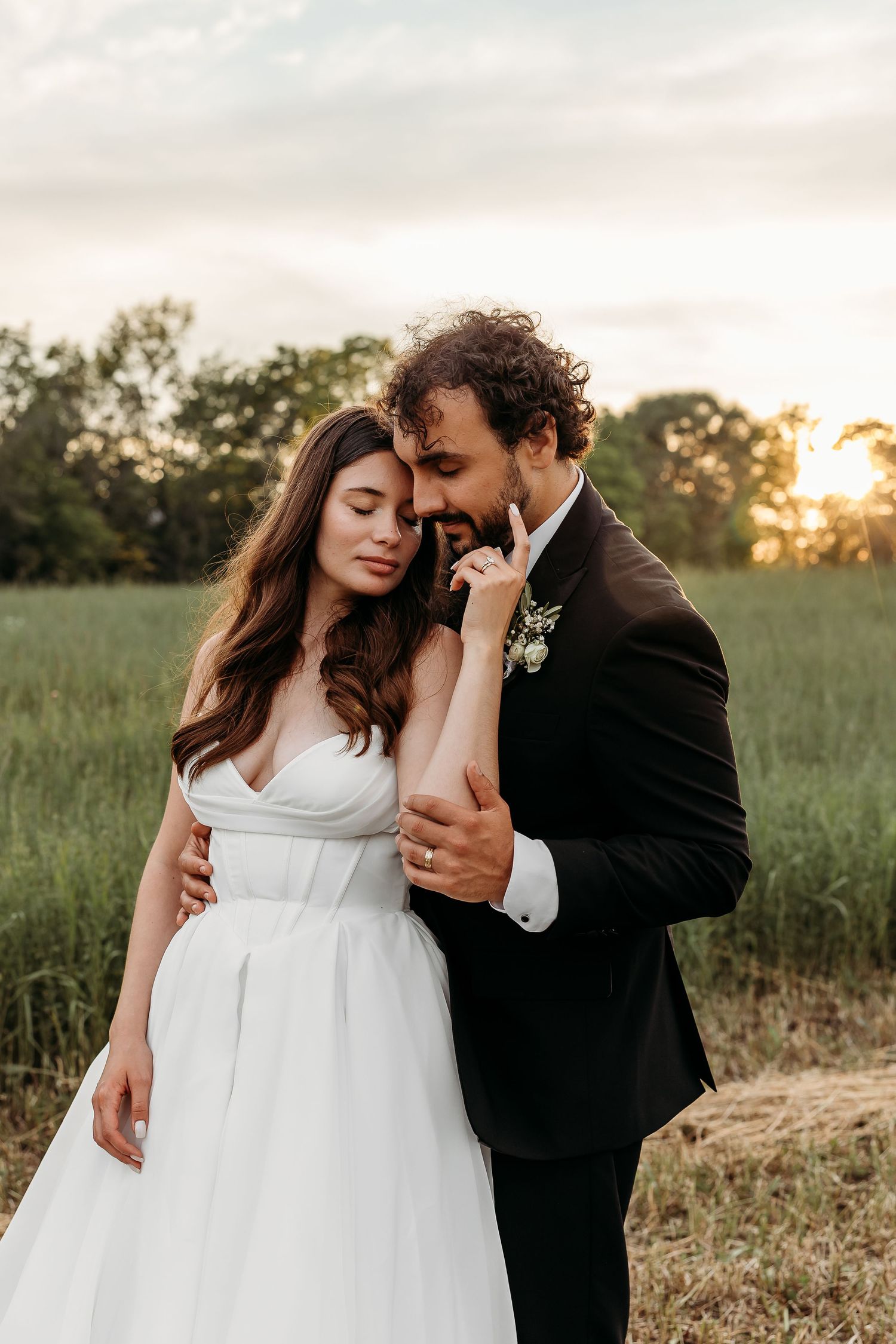 Outdoor wedding photo on grassy field with overcast sky and floral bouquet.