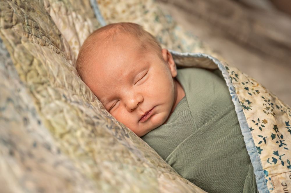 A sleeping newborn baby wrapped in sage green swaddle against floral patterned fabric.