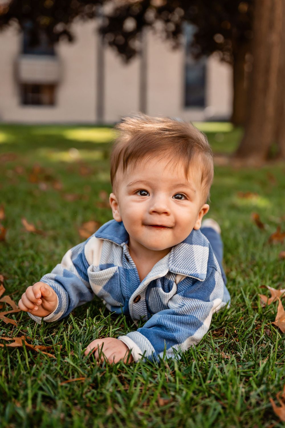 A small child in a blue plaid shirt lies on autumn grass with scattered leaves around them.