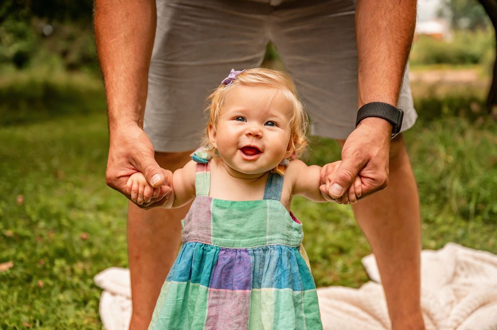 A small child in a colorful dress holds hands with adults while walking on grass and smiling.