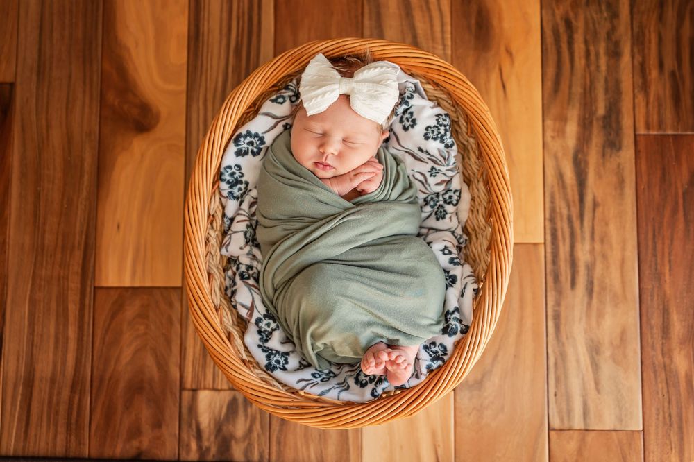 A newborn baby swaddled in sage green fabric lies in a round wooden frame with floral bedding.