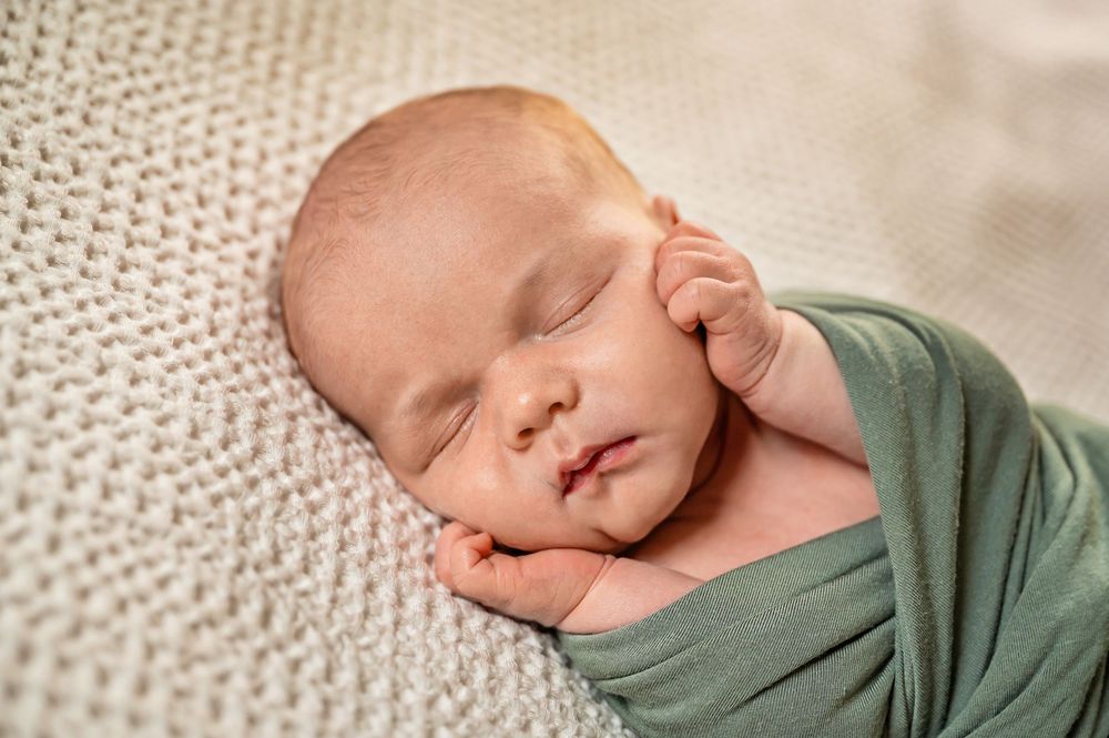 Sleeping newborn baby wrapped in sage green swaddle blanket on textured cream knit backdrop.