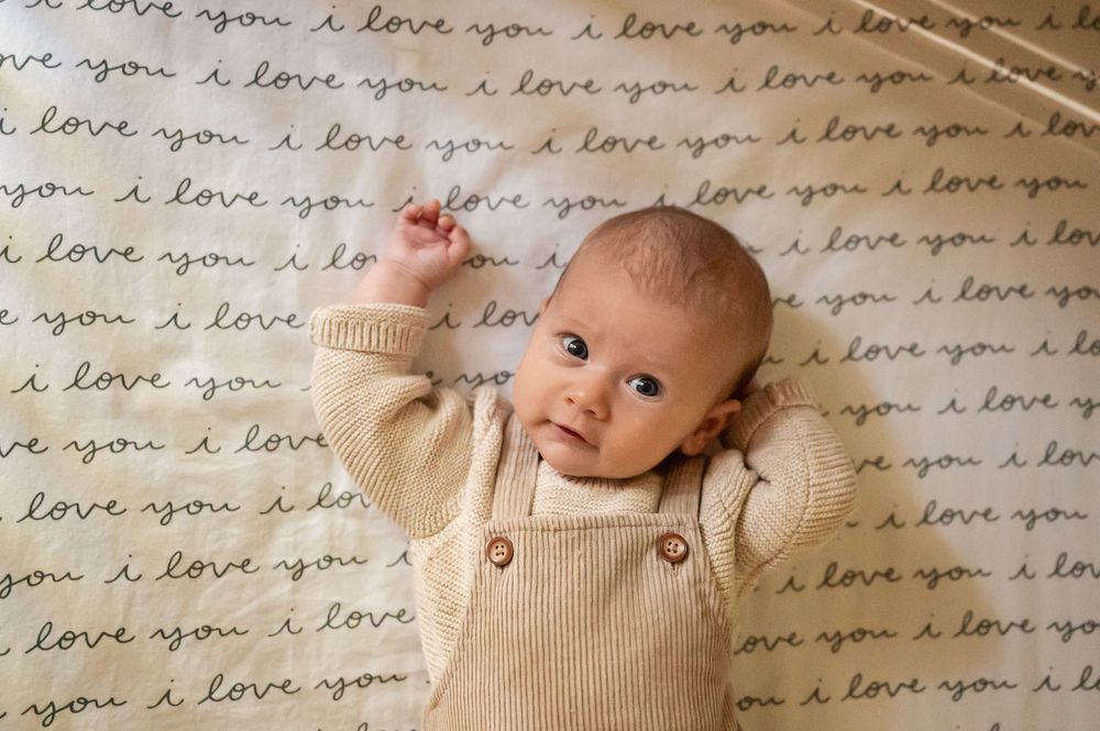 A newborn baby wearing a cozy beige knit sweater lies on a blanket with handwritten 'I love you' text pattern.