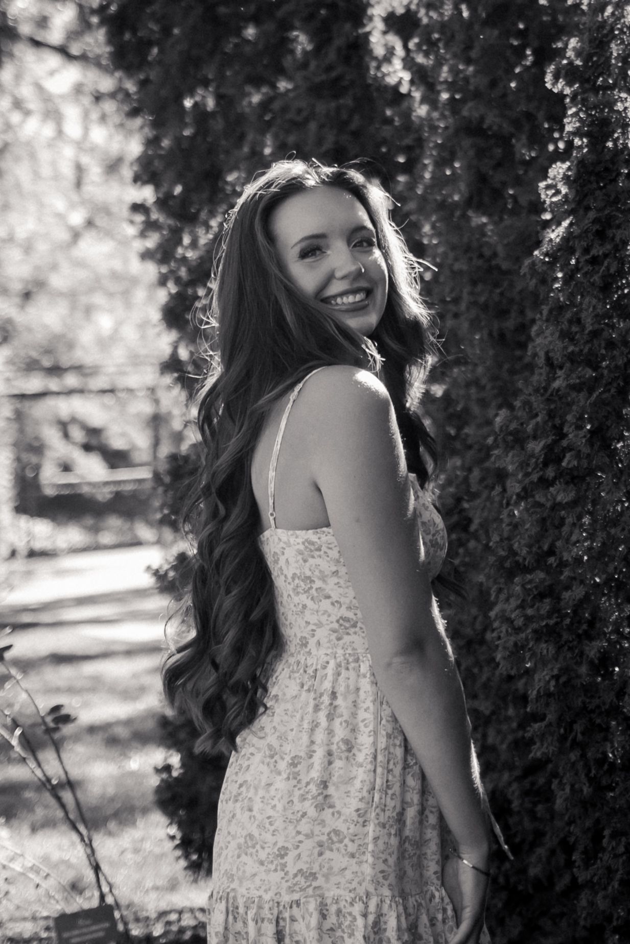 A person in a floral dress smiles while posing against a hedge in black and white photography.