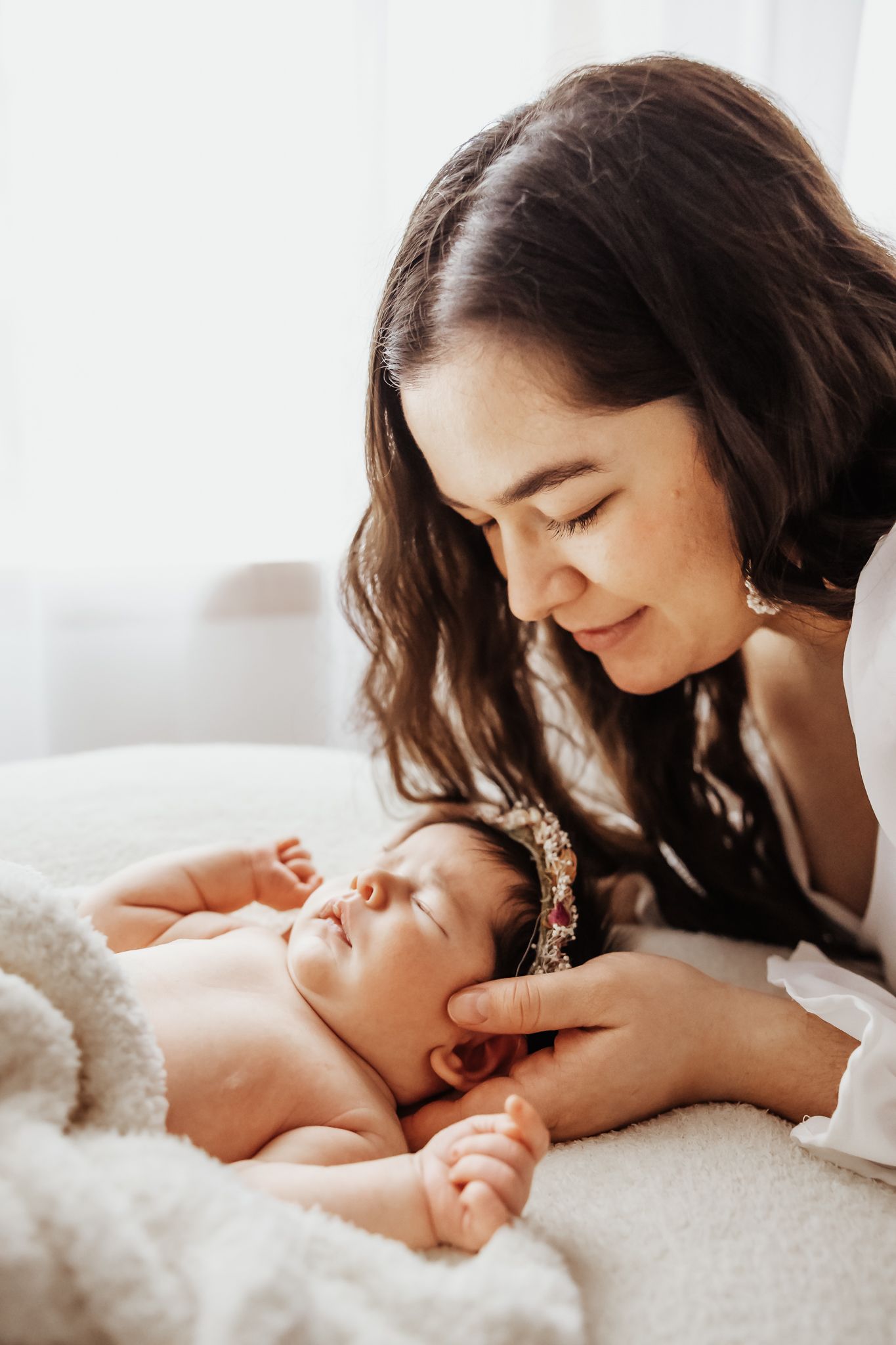 Dark-haired mother gazing lovingly at sleeping newborn baby wearing floral crown on white blanket.