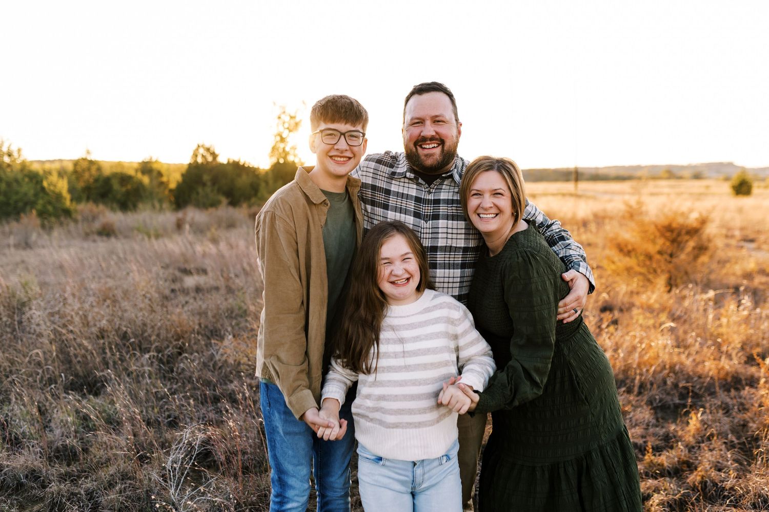Happy family of four posing together in golden field at sunset smiling at camera.