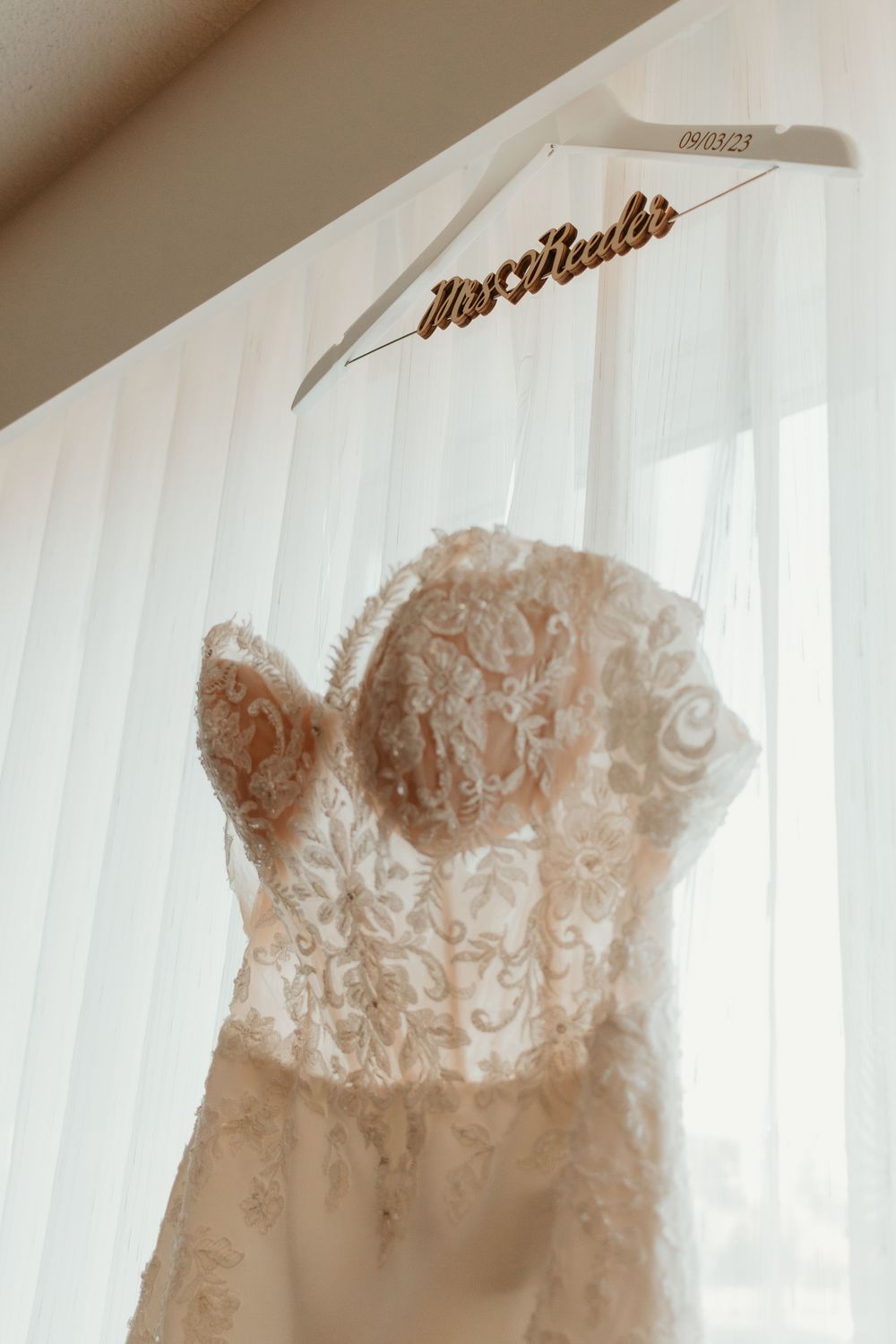 Lace wedding dress hanging on personalized Mrs. hanger against sheer white curtains.