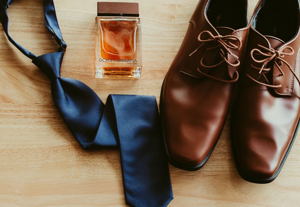 Groom's accessories flat lay featuring brown dress shoes, navy tie, and cologne.