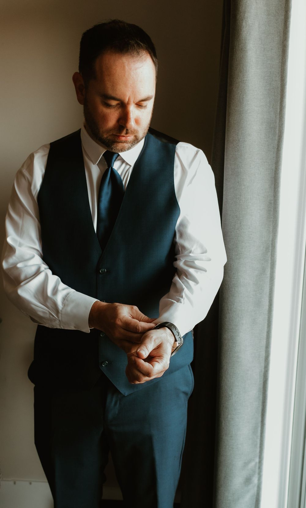 Groom in navy vest and tie adjusting his cufflinks while getting ready for his wedding day.