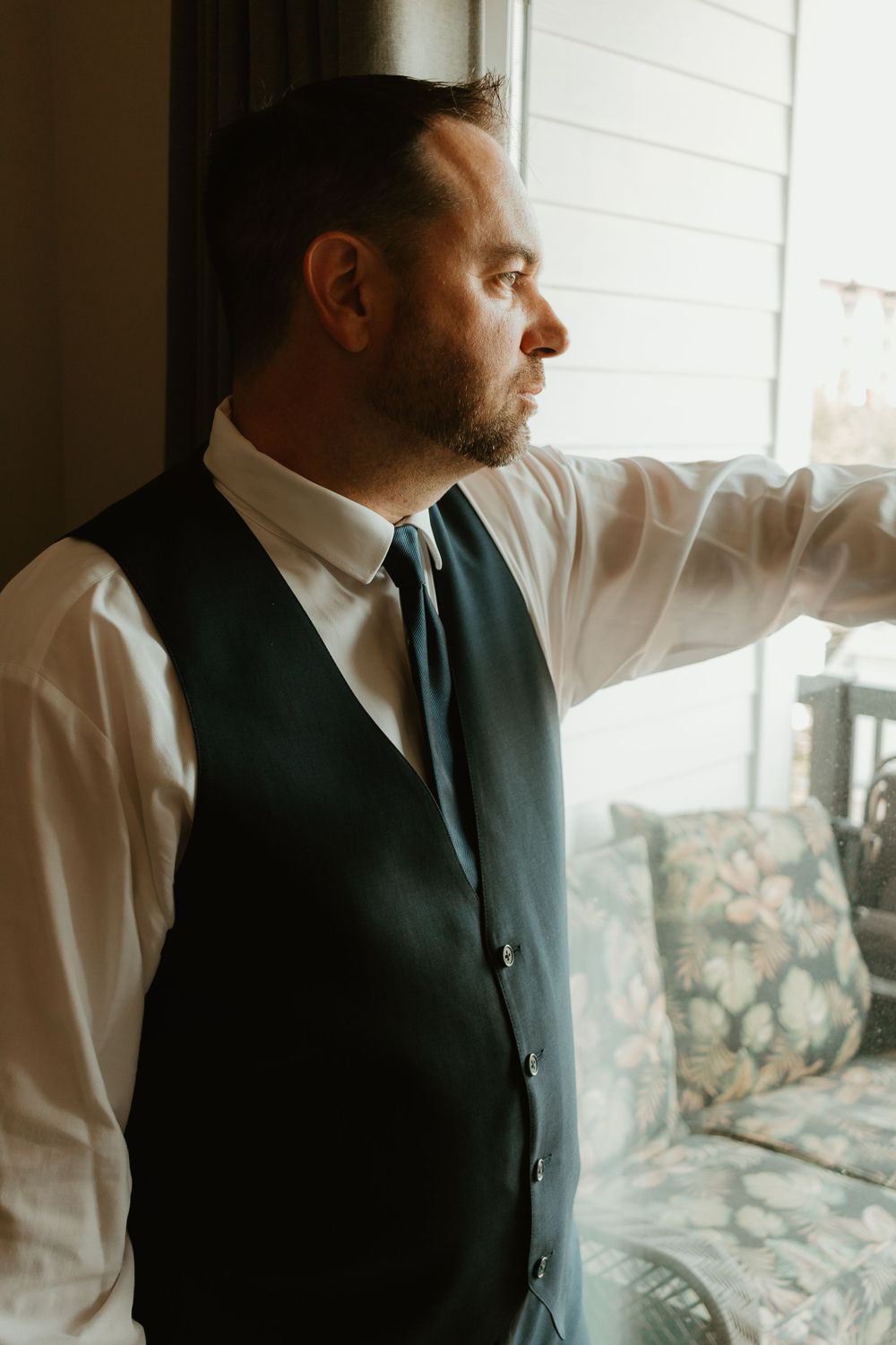 Groom in navy suit vest and tie gazing thoughtfully out a window before his wedding ceremony.