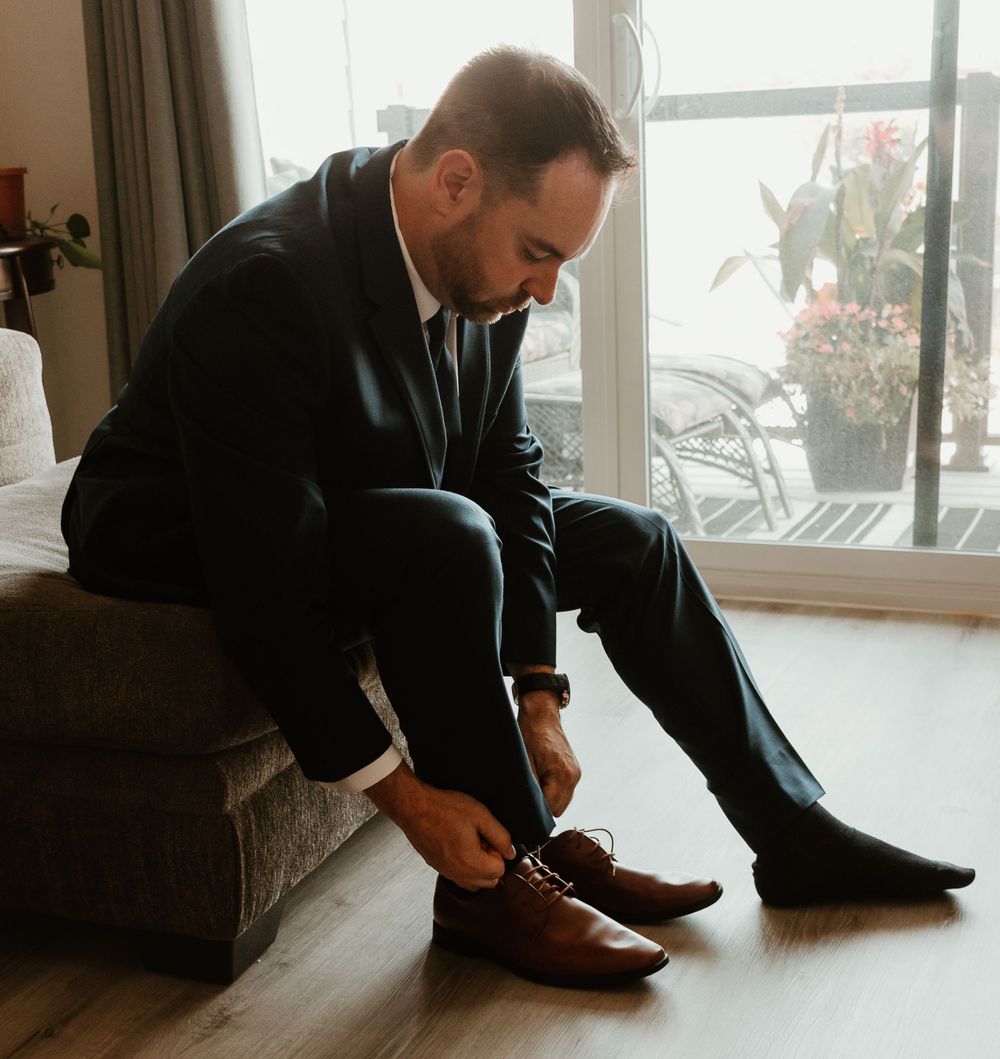 Groom in dark suit tying his brown leather dress shoes while seated on a bench before wedding.