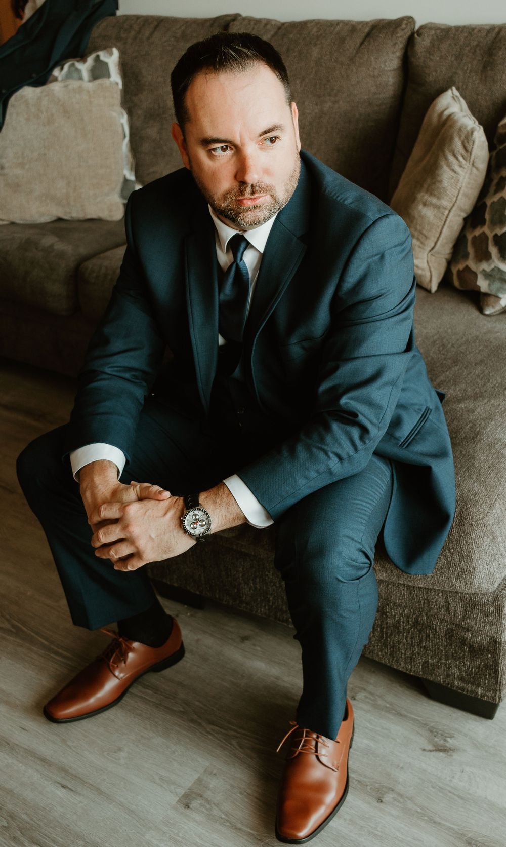 Groom in navy blue suit with brown dress shoes seated on couch posing confidently for wedding photo.