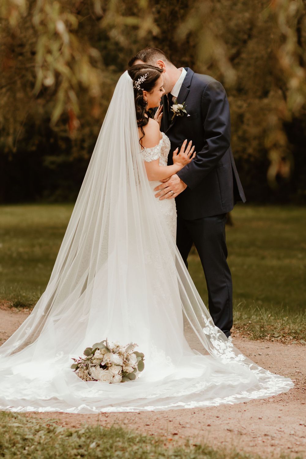 Bride and groom sharing a romantic kiss in lush garden setting with flowing cathedral veil and bouquet.