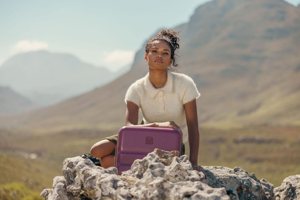 Person sitting on rocky outcrop with purple luggage against mountain backdrop.