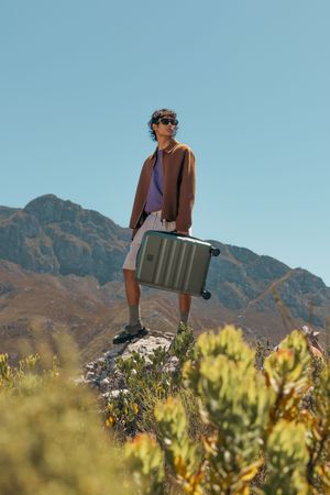 Person standing on rock with grey suitcase surrounded by mountain scenery.