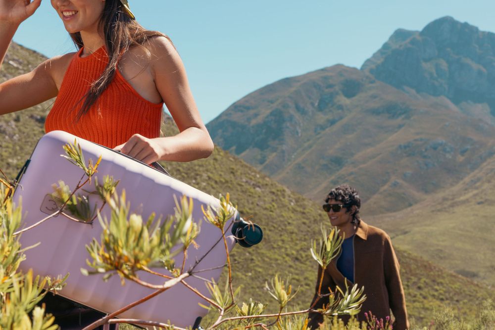 People with luggage exploring sunny mountain terrain with yellow wildflowers.